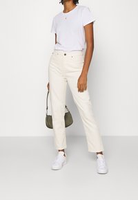 Vit t-shirt, ljusbeiga högmidjade jeans, vita sneakers och en liten olivgrön handväska. Modellen står med ena handen hållandes på väskan.