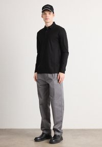 Black long-sleeve polo shirt, gray trousers, and black boots. Model wears a black cap with a white logo, standing on a textured surface.