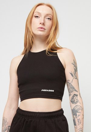 Tânără cu păr roșcat lung poartă un top negru scurt și pantaloni negri, arătând tatuaje cu pasăre, femeie și fluture pe brațul ei stâng.