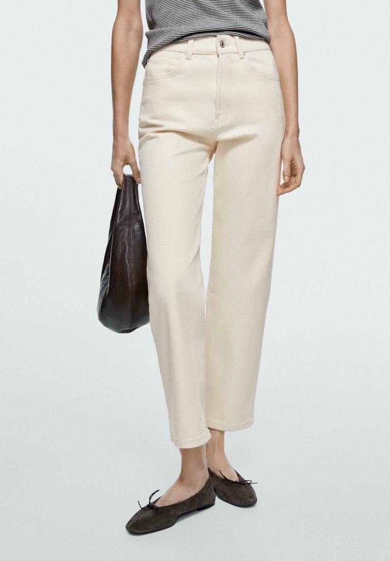 Donna che indossa pantaloni crema a vita alta, top nero a righe, ballerine nere, con una borsa nera in pelle, su uno sfondo chiaro e uniforme.