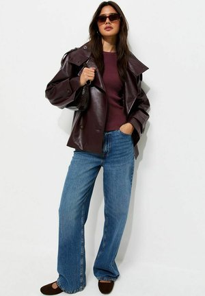 Mujer con chaqueta de cuero marrón oscuro, suéter granate, vaqueros azules, gafas de sol y sujetando un bolso negro sobre fondo blanco.