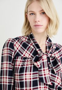 Blusa a quadri con colori blu navy, rosso e grigio. Tessuto morbido con dettaglio al collo a fiocco e tessuto arricciato sotto il busto.