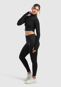 Schwarzes Crop-Top und Leggings aus dehnbarem Material. Lange Ärmel mit Daumenlöchern. Getragen mit weißen Sportschuhen mit schwarzen Akzenten.