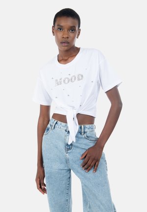 Femme aux cheveux courts portant un crop top blanc noué avec des perles formant le mot « MOOD » et un jean taille haute bleu clair, debout devant un fond blanc.
