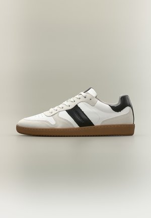 Basket blanche à profil bas avec des détails en daim beige, bande noire sur le côté, patch noir au talon et semelle en caoutchouc marron.
