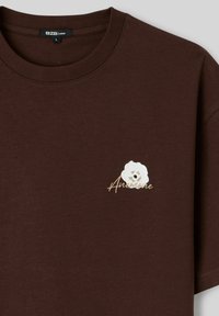 T-shirt ample marron avec une petite fleur blanche et le mot « Anemone » brodé sur le côté gauche de la poitrine.