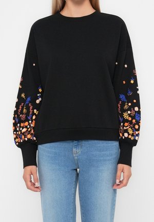 Woman wearing a black sweatshirt with colorful floral embroidery on sleeves paired with light blue jeans, shown from shoulders to hips.