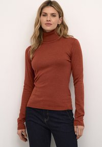Ein rostfarbener Rollkragenpulli aus weichem Material, mit einer figurbetonten Passform und gerippten Bündchen, kombiniert mit dunklen Jeans für einen Kontrast.
