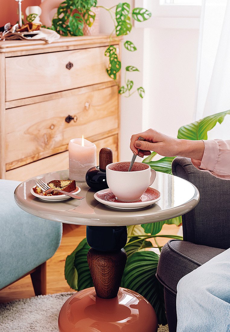 Runder Tisch mit einer glänzenden grauen Oberfläche, Holzfuß und korallenfarbenem Sockel. Auf ihm stehen eine rosa Tasse und Untertasse, ein Dessert und eine Kerze.