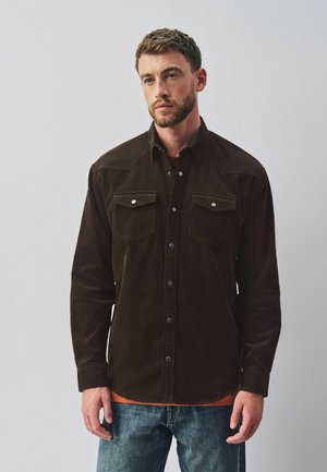 Man with short hair and beard wearing a dark brown button-up shirt over an orange shirt and blue jeans, standing against a light background.
