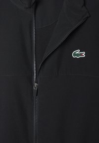 Zwarte zip-up jack met gedeeltelijk open rits, voorzien van een klein groen en rood krokodillenlogo op de borst.