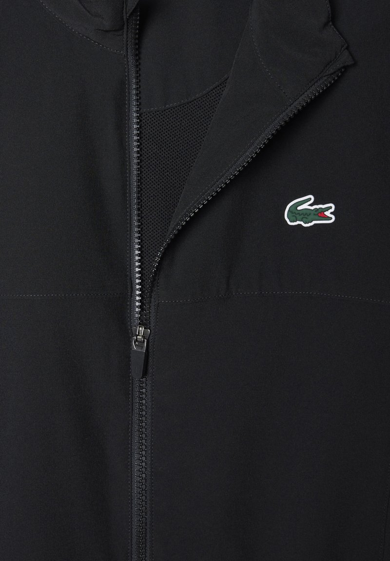 Schwarze Zip-Jacke mit teilweise geöffnetem Reißverschluss, mit einem kleinen grünen und roten Krokodil-Logo auf der Brust.