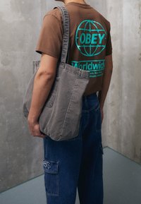 Šedá plátěná taška s dvojitě šitými stranami, nošená přes rameno, v kombinaci s modrými džíny cargo s vyšívanými detaily.