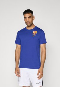 Plava pamučna majica s okruglim izrezom, sadrži žuti Nike logo i višebojni grb FC Barcelone na lijevoj strani prsnog koša.