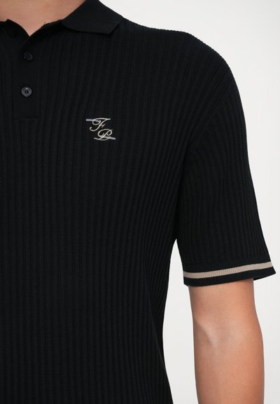 Detailný záber na osobu s čiernym rebrovaným polo tričkom s krátkym rukávom a béžovo pruhovaným manžetovým pásikom, na hrudi vyšívané logo "FP".
