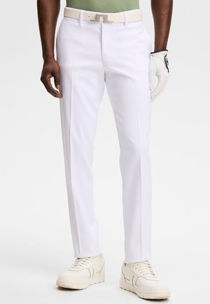 Homme portant un pantalon blanc sur mesure, une ceinture blanche, un gant de golf blanc à la main droite, des baskets beige, et une chemise verte rentrée.