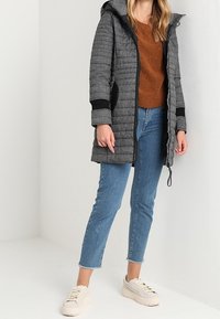 Gequilted grijs- en zwartgekleurde hoodie met ritssluiting, zijzakken en elastische manchetten, gecombineerd met een bruine trui en blauwe spijkerbroek.