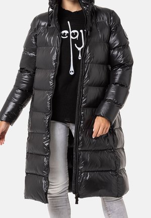 Persoon draagt een lange, glanzende zwarte donsjas met capuchon, over een zwarte trui met wit grafisch ontwerp en lichtgrijze strakke spijkerbroek.