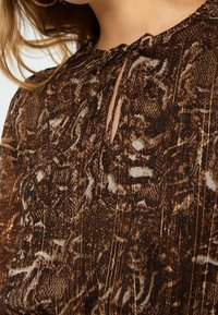 Brun blus med orm-lädermönster, med fina veck, en knytkrage-design och metalltrådsinslag för extra textur.