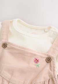 Cream long-sleeve baby shirt under light pink corduroy dress with embroidered pink flower and wooden buttons on straps.