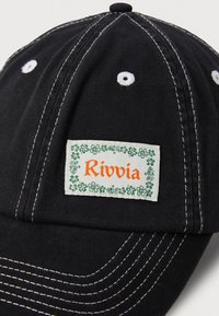 Casquette de baseball noire en coton avec des coutures blanches contrastantes ; présente un patch rectangulaire du logo Rivvia entouré de détails floraux verts.