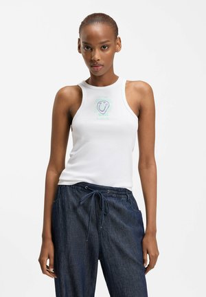 Femme mince portant un haut blanc sans manches avec un motif de visage souriant et un pantalon bleu foncé à cordon, debout devant un fond uni.