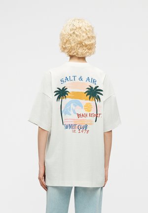 Osoba o kręconych blond włosach, ubrana w białą, oversize'ową koszulkę z kolorową grafiką ośrodka nadmorskiego i tekstem na plecach.