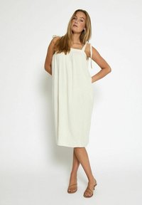 Frau in einem ärmellosen cremefarbenen Midikleid mit Bindeschlaufen und nudefarbenen Absatzsandalen, die mit einer Hand auf der Hüfte vor einem weißen Hintergrund posiert.