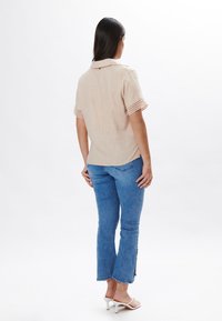 Camisa corta de manga corta color beige con cuello y adornos de encaje, combinada con jeans de mezclilla de lavado claro con extremos acampanados y aberturas laterales.