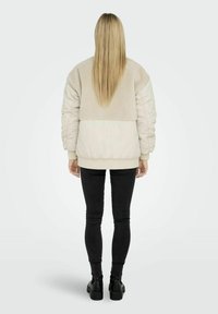 Giacca beige foderata in pile con maniche voluminosi, una parte inferiore liscia, polsini a coste e vita elastica, abbinata a jeans skinny neri.