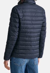 Person, der eine schwarze gesteppte Jacke und blaue Jeans trägt, von hinten vor einem einfarbigen weißen Hintergrund gezeigt.