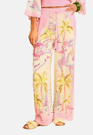 Lyserøde bukser med vidde i benene med tropisk print af palmer, flamingoer, sejlbåde og strandscener, båret med flettede sandaler og et grønt armbånd.