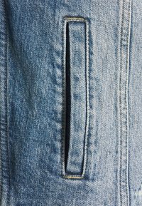 Ljusa blå denimtyg med en subtil textur, med en enda rektangulär ficka på sidan, omgiven av kontrasterande sömnad.