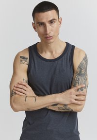 Dunkelgraues ärmelloses Tanktop mit einem kontrastierenden marineblauen Halsausschnitt. Es hat einen figurbetonten Schnitt und sichtbare Tattoos an den Armen.