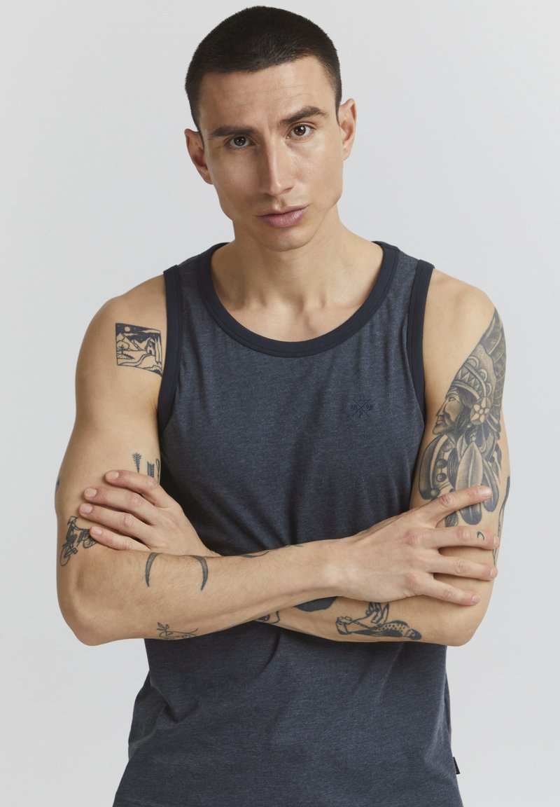 Dunkelgraues ärmelloses Tanktop mit einem kontrastierenden marineblauen Halsausschnitt. Es hat einen figurbetonten Schnitt und sichtbare Tattoos an den Armen.
