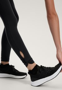 Fekete leggings, bokai hasítékokkal, fekete sportcipővel párosítva, amelynek textúrázott felsőrésze és fehér talpa van. Simára finomított, funkcionális dizájn.