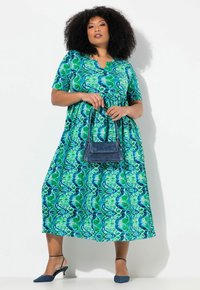Grünes und blaues gemustertes Maxikleid mit kurzen Ärmeln und V-Ausschnitt. Kombiniert mit einer dunkelblauen Handtasche und passenden blauen High Heels.