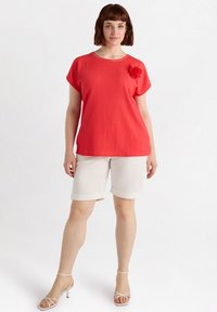 Top rojo de manga corta con tejido texturizado y un acento floral, combinado con pantalones cortos beige claros de corte tailored y tacones blancos con tiras.