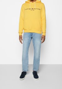 Sudadera amarilla con bolsillo frontal y cordones, con el logo negro "TOMMY HILFIGER EST. 1985". Combinada con vaqueros azul claro y zapatillas náuticas azul marino.