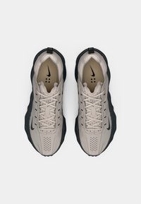 Sapatilhas Nike de tom bege claro apresentam um upper texturizado, sola de borracha preta e um sistema de atacadores proeminente. O design inclui detalhes perfurados e um logo.