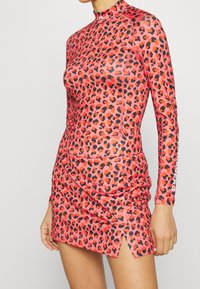 Long-sleeve, high-neck dress in a pink fabric with a black and red leopard print. Features ruching and side slits for movement.