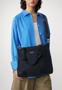 Svart canvas tote-väska med två handtag och ett avtagbart rem. Har en präglad logotyp och en jämn yta. Matchad med en blårandig skjorta.