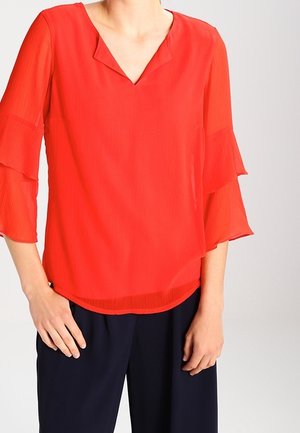 Femme portant un chemisier rouge vif à manches longues avec des manches à volants et un col en V, associé à un pantalon bleu marine foncé sur un fond uni.