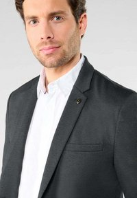 Blazer gris texturé avec un revers cranté, une poche poitrine simple et un petit logo en accent. Porté par-dessus une chemise blanche à col.