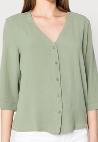Blusa verde chiaro, con bottoni e scollo a V, a maniche tre quarti. Tessuto texturizzato e finitura morbida. Sei bottoni frontalmente.