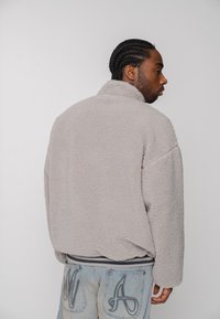 Mand med fletninger iført en grå fleecejakke og lyseblå jeans med broderede lommer, set i profil mod en ensfarvet baggrund.