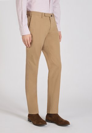 Homme portant un chino beige associé à des bottines Chelsea en daim marron et une chemise rayée rose clair, debout devant un fond uni.
