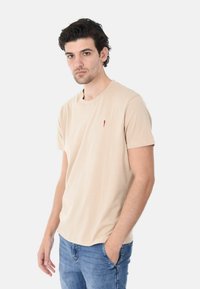 T-shirt beige a maniche corte realizzato in un tessuto morbido. Presenta un piccolo accento rosso sul petto sinistro. Abbinato a jeans blu. Design semplice e casual.
