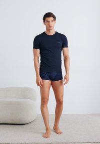 T-shirt noire ajustée à manches courtes, en tissu doux, associée à un boxer en coton bleu marine. Le mannequin se tient sur un tapis clair à côté d'une chaise crême arrondie.