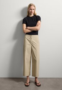Schwarzes, eng anliegendes Kurzarmoberteil kombiniert mit beigen, weit geschnittenen Hosen und schwarzen Flip-Flops. Glattes Material, schlichtes Design und neutrale Farben.